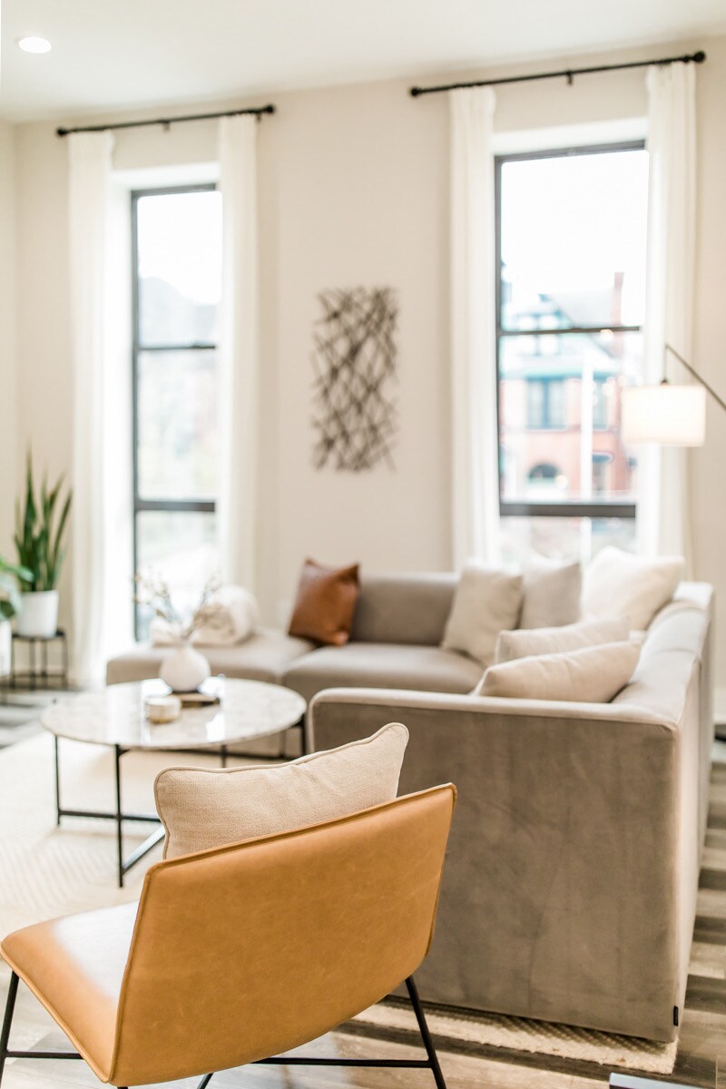 A modern living area features a sectional sofa adorned with light-colored cushions, positioned near two tall windows that allow natural light. A round coffee table occupies the center, accompanied by a sleek armchair in a warm leather tone. Decorative plants and wall art add subtle elegance.