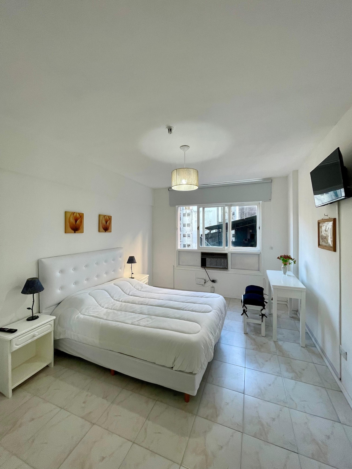 A well-lit room features a spacious bed with a white headboard, flanked by matching nightstands and lamps. A small desk is positioned near a window, which offers a view of the cityscape. A television is mounted on the wall opposite the bed.