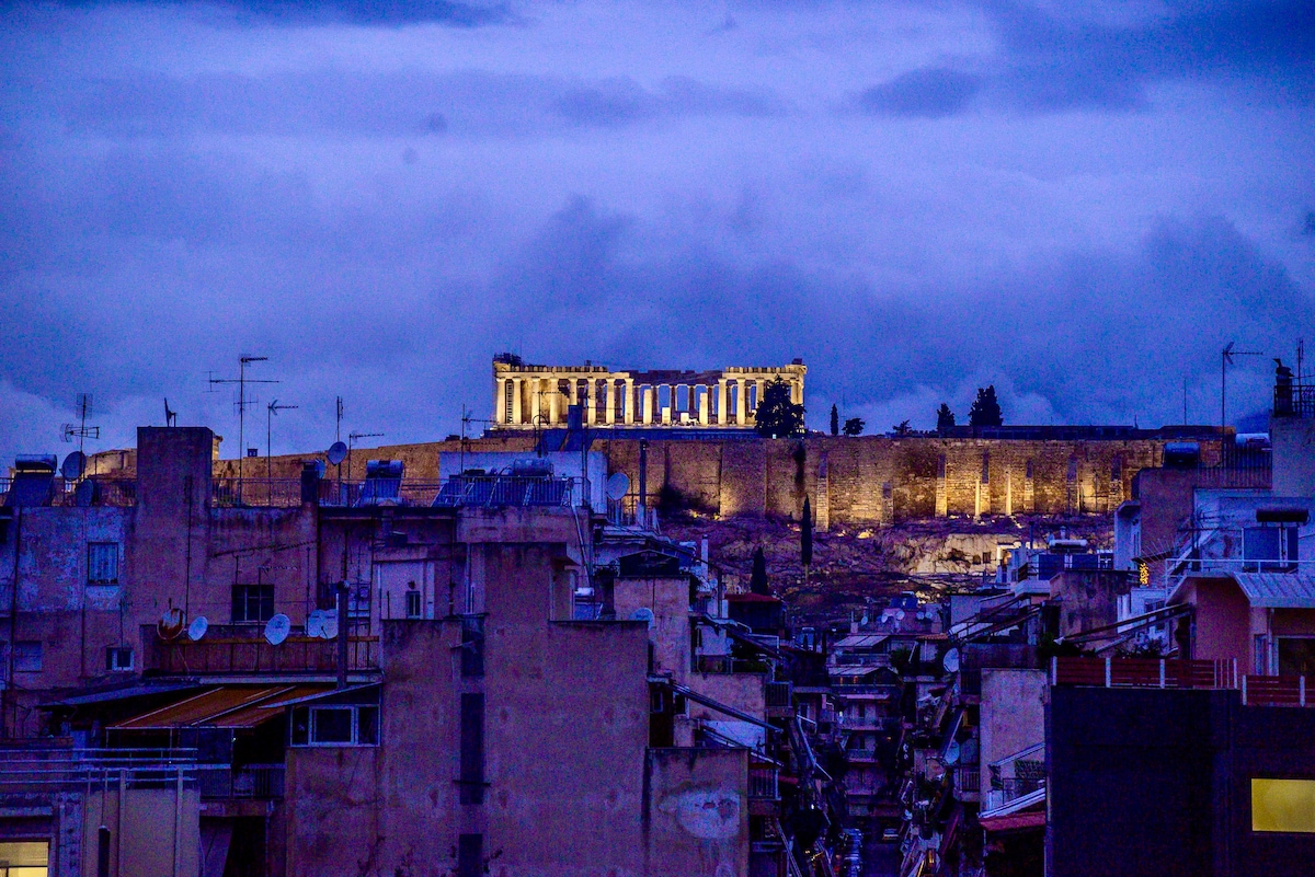 The Acropolis stands majestically illuminated against a twilight sky, showcasing ancient architecture. The surrounding urban landscape features a mix of rooftops and structures, with scattered antennas and balconies visible. The scene reflects a contrast between the historic site and the modern city.
