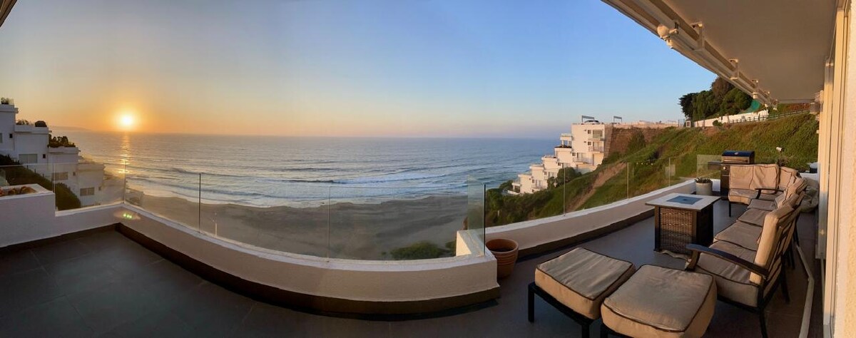 A panoramic view of the ocean at sunset is displayed from a spacious terrace. Comfortable lounge furniture is arranged for relaxation, with glass barriers providing unobstructed sightlines to the horizon. The coastline is visible in the background, complementing the tranquil seaside setting.