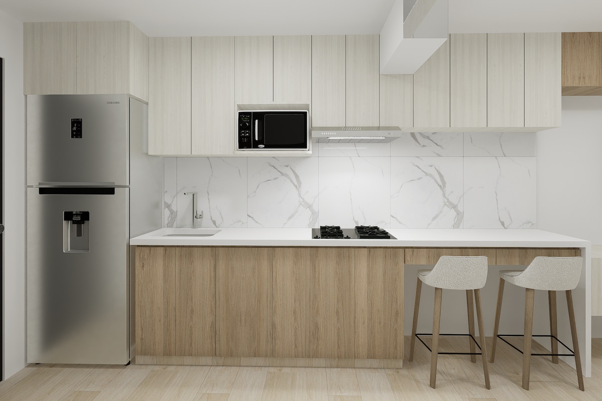A modern kitchen area features cabinetry with a light wood finish and white countertop. Stainless steel appliances, including a refrigerator and microwave, are integrated. Two stools are positioned at the breakfast bar, enhancing the functionality of the space.