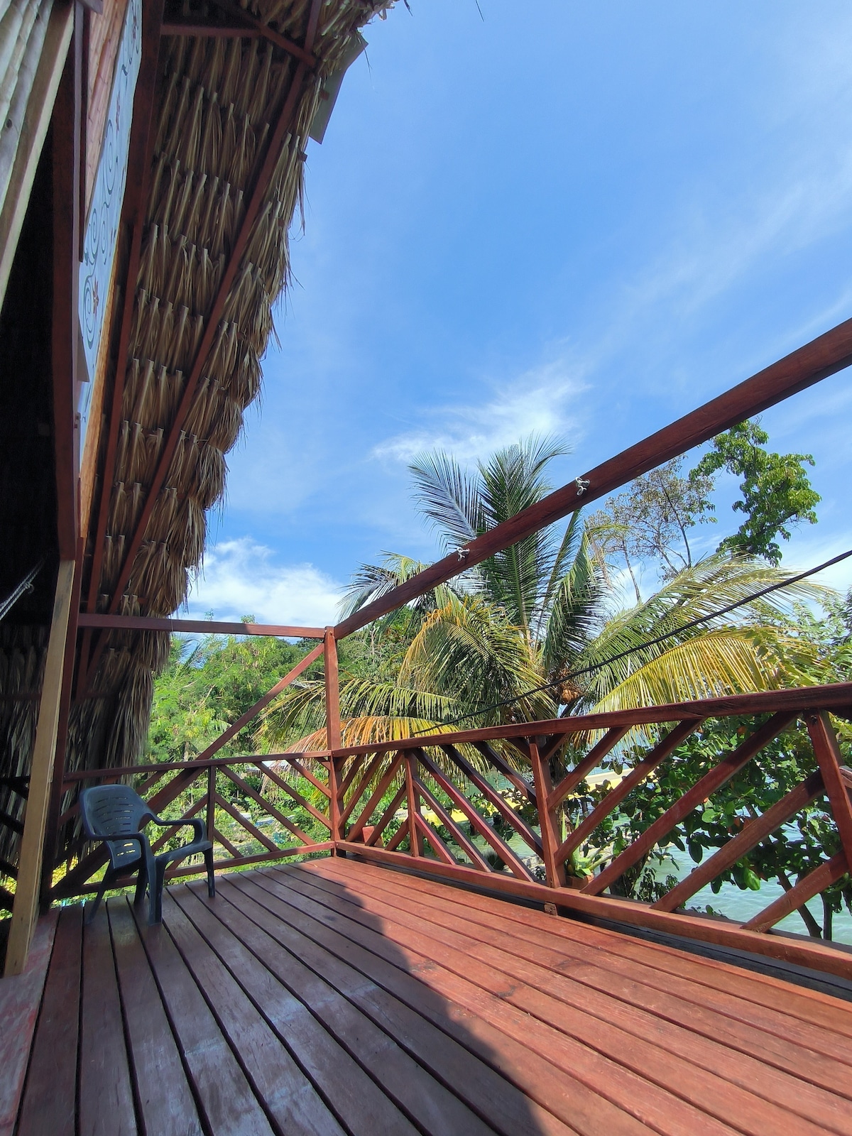 A spacious wooden deck is featured, surrounded by lush greenery and accented by tall palm trees. The structure is adorned with a thatched roof, and a single chair is positioned thoughtfully on the deck, offering an inviting space to enjoy the natural surroundings.