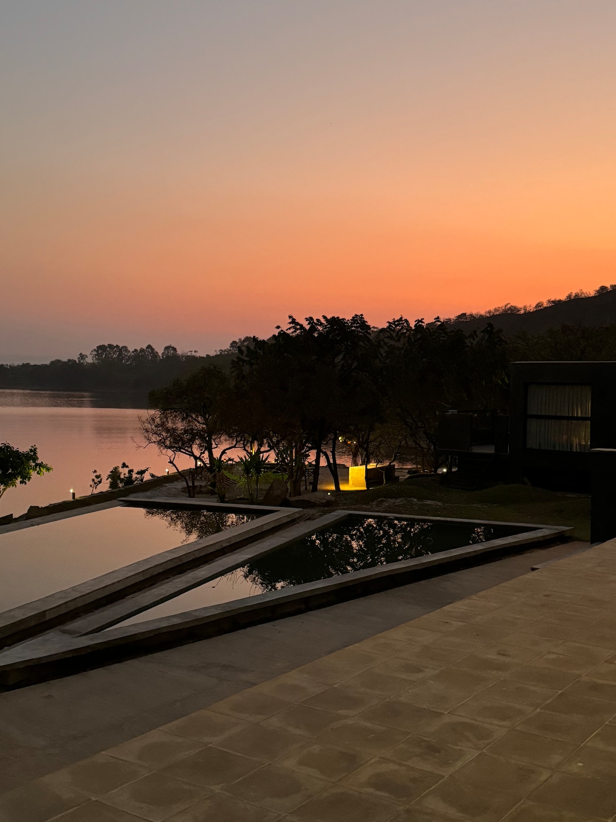 A serene view at dusk showcases tranquil water reflecting the colorful sky. Trees line the shore, with soft illumination from nearby lights enhancing the peaceful ambiance. The image captures a modern structure with clean lines and large windows overlooking the scenic landscape.