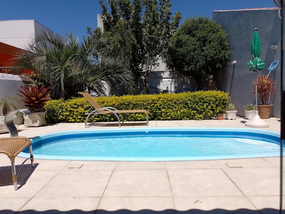 A private pool area is presented with clear blue water, surrounded by lush greenery and various potted plants. A lounge chair is positioned beside the pool, while a shaded area provides a comfortable spot to relax.