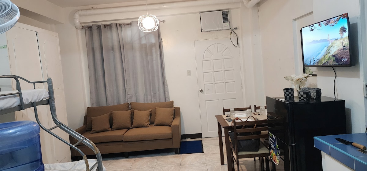 The living area is equipped with a brown sofa facing a wall-mounted television. A dining table with four chairs sits adjacent to a black cabinet. Natural light filters through sheer curtains, while an air conditioning unit is positioned above the entrance door.