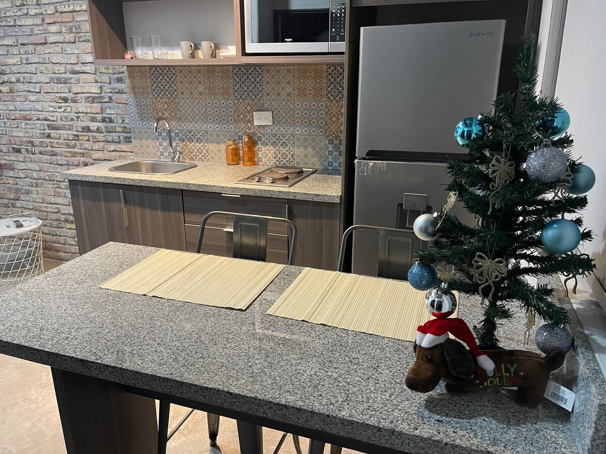 A kitchen space features granite countertops complemented by a modern sink and appliances. Decorative tiles add visual interest to the backsplash. A small holiday tree with blue ornaments sits on the table, which is set with two placemats. Soft lighting enhances the cozy ambiance.