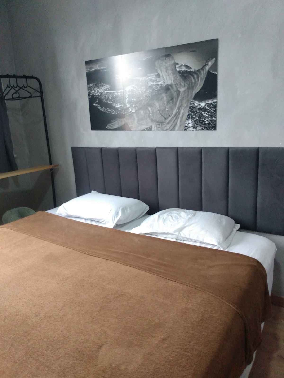 A spacious bed is outfitted with white pillows and a plush brown blanket, creating a comfortable sleeping area. A wall-mounted artwork featuring the Christ the Redeemer statue is displayed above the headboard, enhancing the room's overall aesthetic.