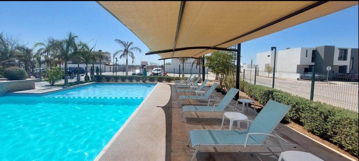 A serene pool area is shown, featuring a sparkling blue swimming pool surrounded by lounge chairs under shaded sun umbrellas. Lush greenery lines the perimeter, with palm trees adding a touch of nature. The gated community is visible in the background.
