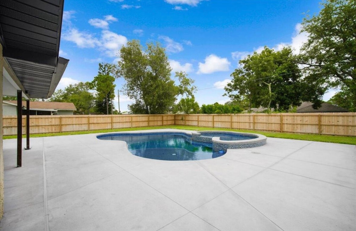 The backyard features a newly built lagoon-style pool, surrounded by a spacious patio area. A wooden fence encloses the space, providing privacy. Lush greenery is visible beyond the fence, contributing to a tranquil outdoor environment.