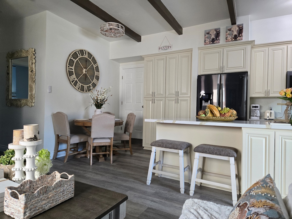 The open-concept kitchen and dining area showcases light-colored cabinetry and modern appliances. A round dining table is accompanied by upholstered chairs, while a large clock adorns the wall. Bar stools are positioned at the kitchen island, inviting casual dining or gathering space.