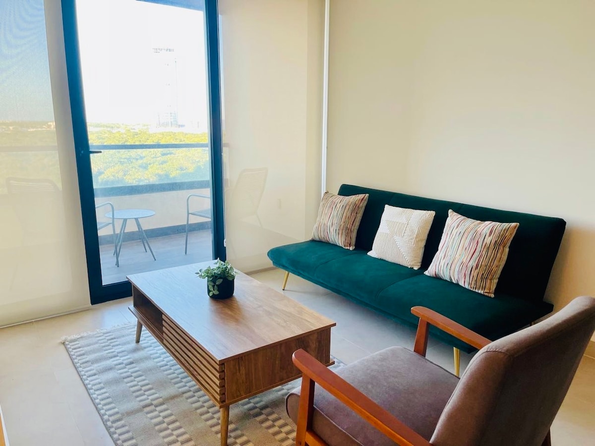 A light-filled living area features a green sofa adorned with patterned cushions. A wooden coffee table sits centrally, accompanied by a cozy armchair. Visible through large glass doors is a balcony with outdoor seating, offering a view of greenery.