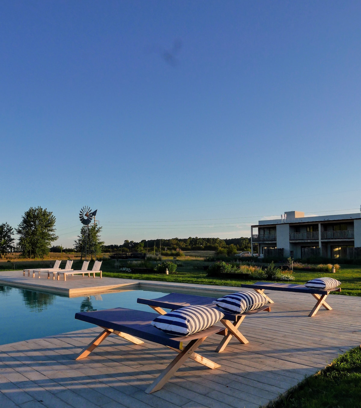 The outdoor area features a refreshing pool surrounded by wooden decking and lounge chairs adorned with striped cushions. Nearby, a windmill adds character to the scenic landscape, while a modern two-story building is visible in the background under a clear blue sky.