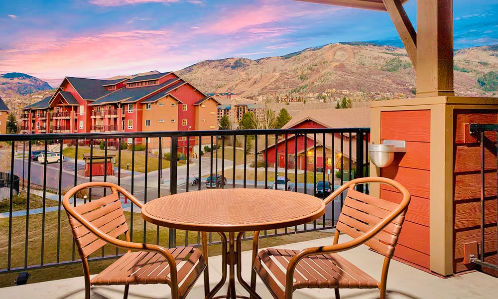 A private balcony features a round table with two woven chairs, offering scenic views of the surrounding mountains and neighboring buildings. The sky is painted with soft hues of pink and lavender during sunset, creating a tranquil outdoor setting.