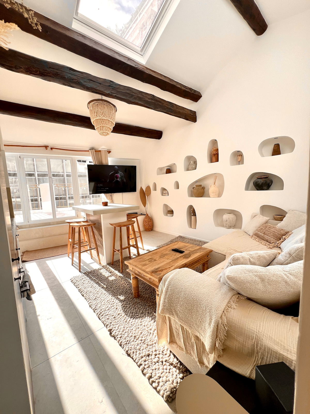 A bright and airy living space features high ceilings with wooden beams and a large window allowing natural light. Shelves with decorative items line the walls, and a cozy seating area includes a wooden coffee table and comfortable couches, creating a relaxed atmosphere.