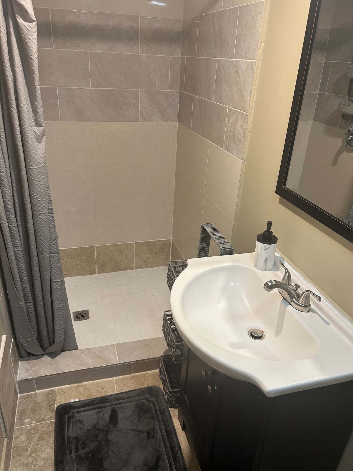 A modern bathroom features a tiled shower area with a sleek, glass partition. An elegant sink is positioned on a dark vanity, accompanied by a stylish mirror above. A soft gray bath mat lies on the floor, adding a touch of comfort to the space.