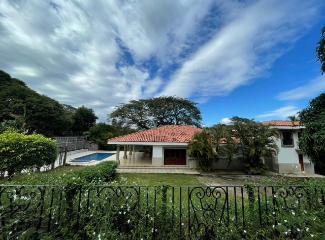 A spacious single-story house with a red-tiled roof is surrounded by lush greenery. A large swimming pool is visible in the backyard, while a decorative wrought-iron fence frames the front garden. The sky features scattered clouds, enhancing the outdoor setting.