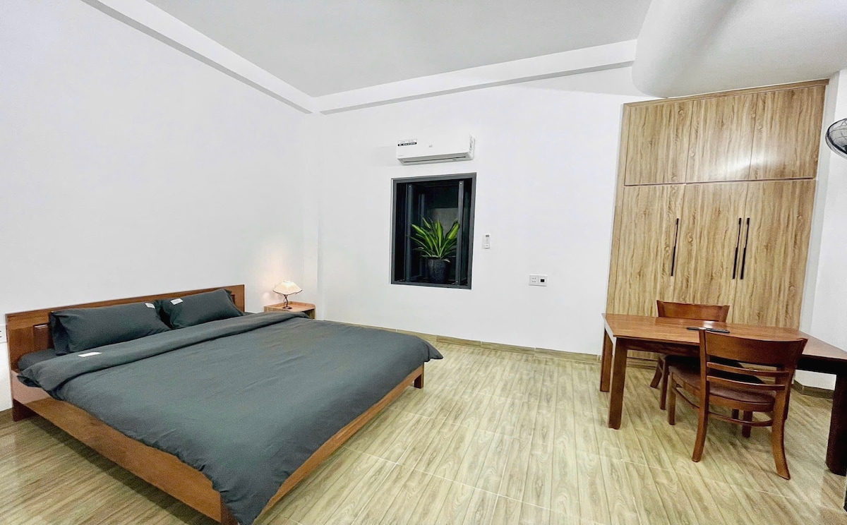 A modern bedroom features a spacious bed with a dark grey coverlet. A small bedside lamp provides soft illumination. A dining table with two chairs sits against the wall, while a large window allows natural light to enter, surrounded by light-colored walls and wooden flooring.