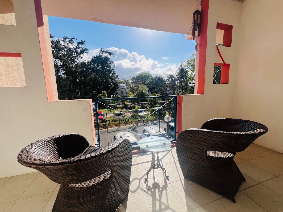 A small private balcony is presented, featuring two woven chairs positioned around a circular table. Sunlight illuminates the space, with a view of the surrounding greenery and the road. The balcony's walls are accented in warm tones, providing a cozy outdoor setting.
