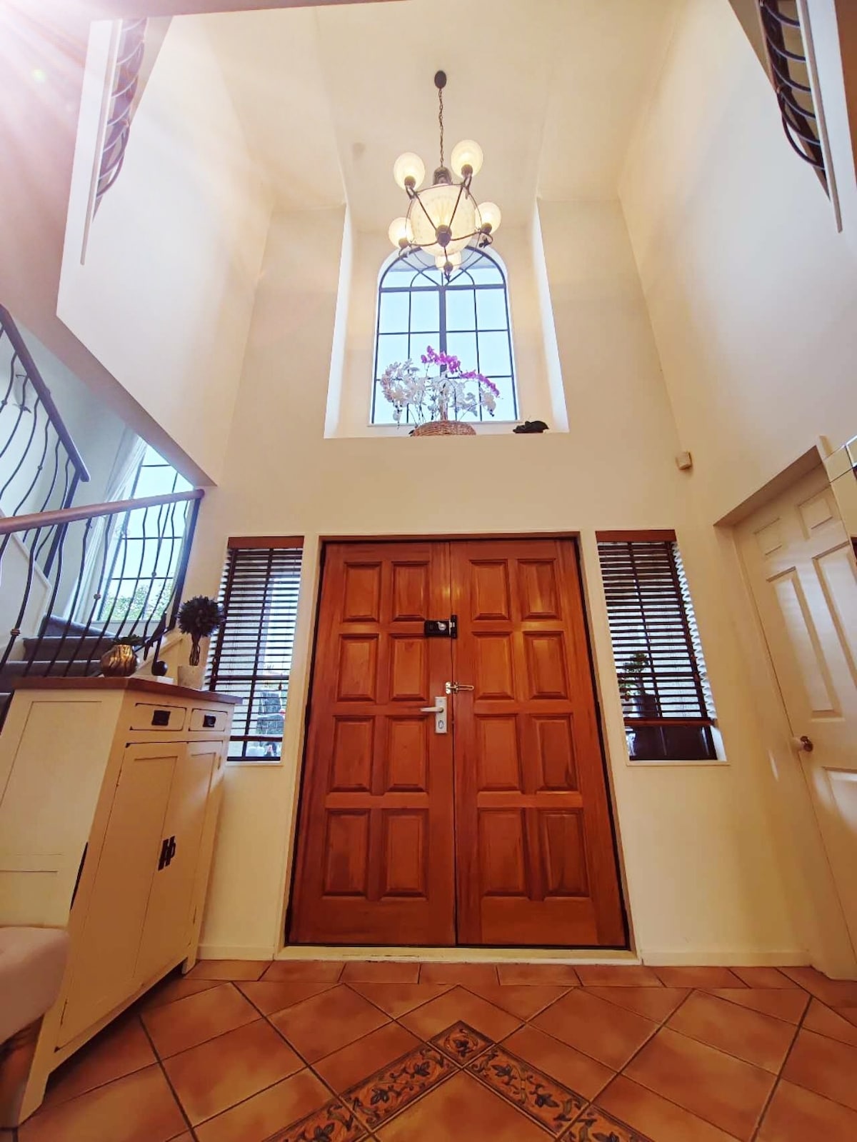 The entrance features a tall ceiling with a chandelier creating a bright and airy atmosphere. A large wooden door is visible, complemented by sidelights and elegant window treatments. A decorative piece rests on a console table, enhancing the welcoming entryway.