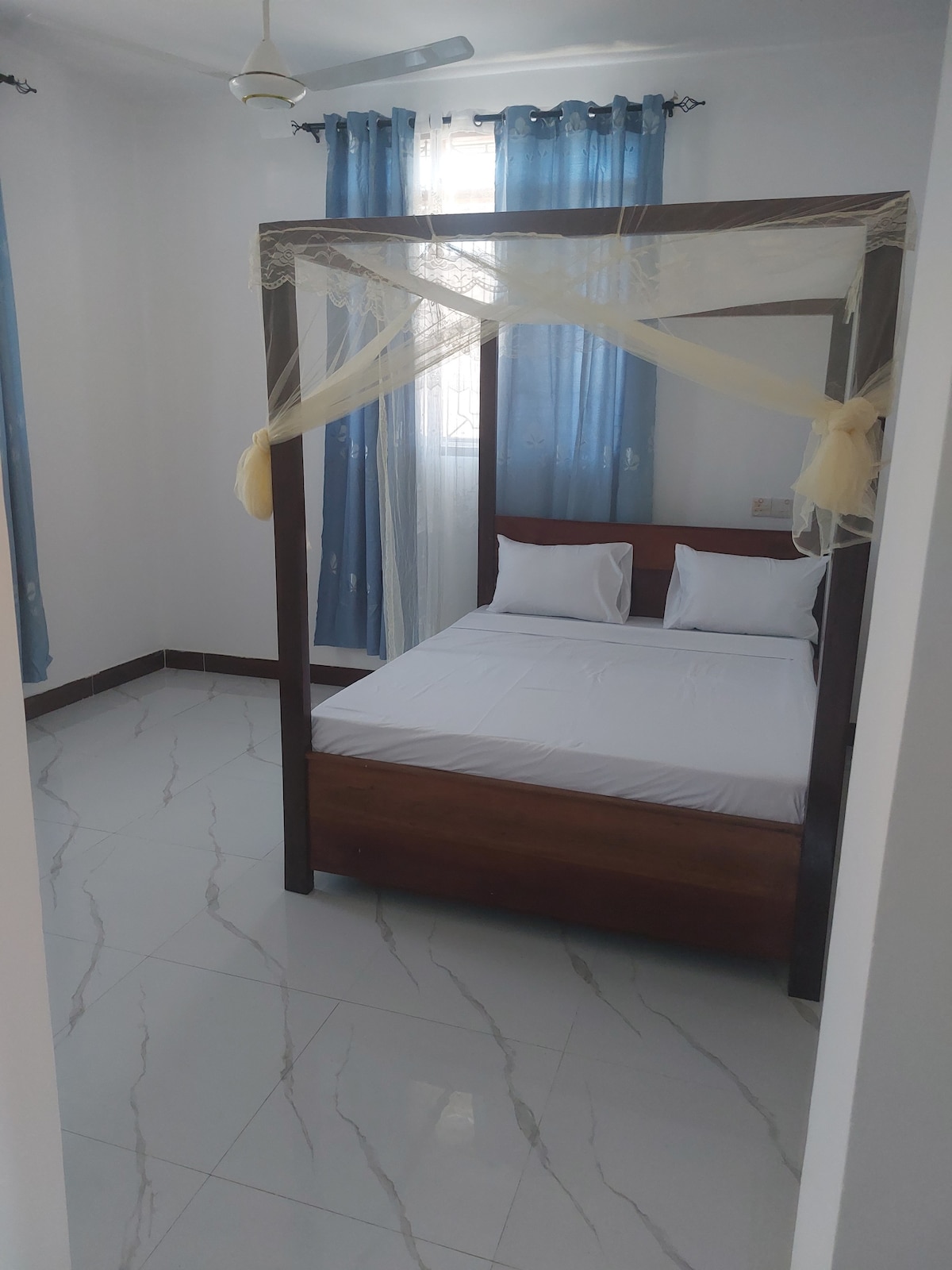 A well-lit bedroom features a four-poster bed draped with light fabric. Soft natural light filters through the sheer blue curtains. The tiled floor adds a clean touch, and two white pillows are arranged neatly on the bed.