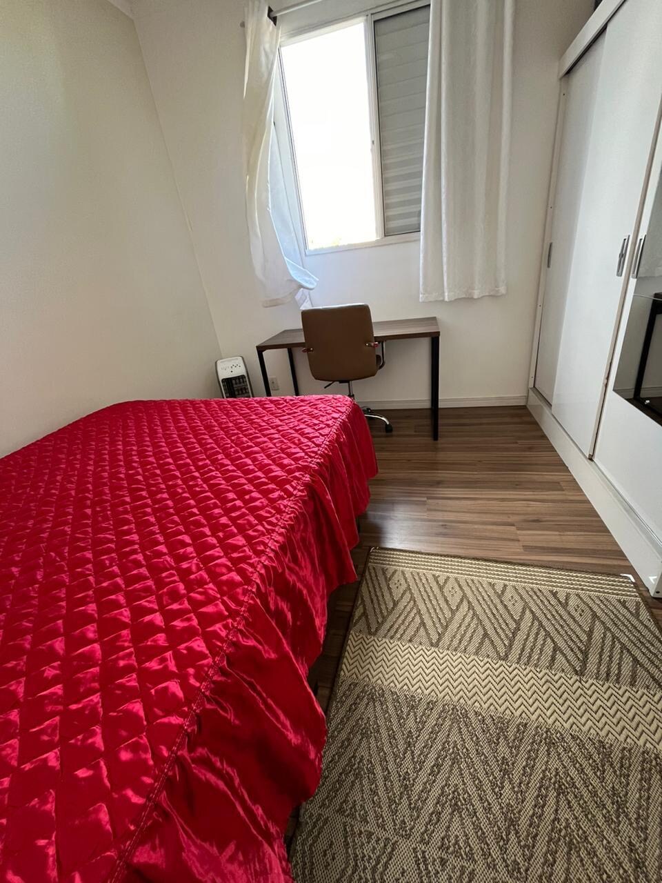 A cozy sleeping area features a red quilted bedspread atop a spacious bed. A chair and desk are positioned nearby, with natural light entering through the window covered by sheer curtains. A patterned area rug adds warmth to the hardwood floor.