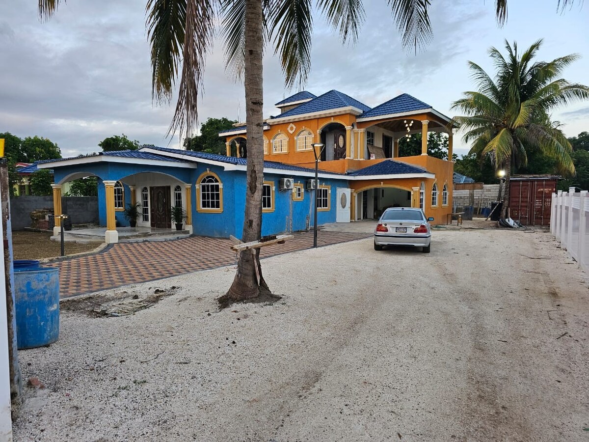 A brightly colored two-story house features a blue roof and yellow exterior walls. The driveway leads to the entrance, with palm trees framing the property. A gravel area is visible in the foreground, along with a glimpse of lush greenery beyond the house.