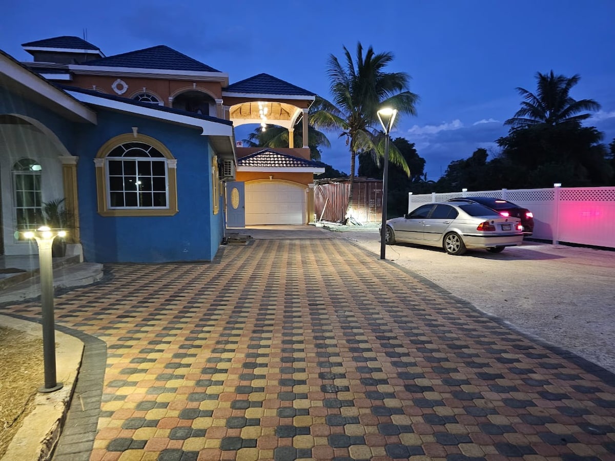The exterior of the property is captured at dusk, showcasing a paved driveway leading to a garage. The driveway features colorful stones, illuminated by pathway lights. A car is parked on the right side, with palm trees and a gazebo visible in the background.