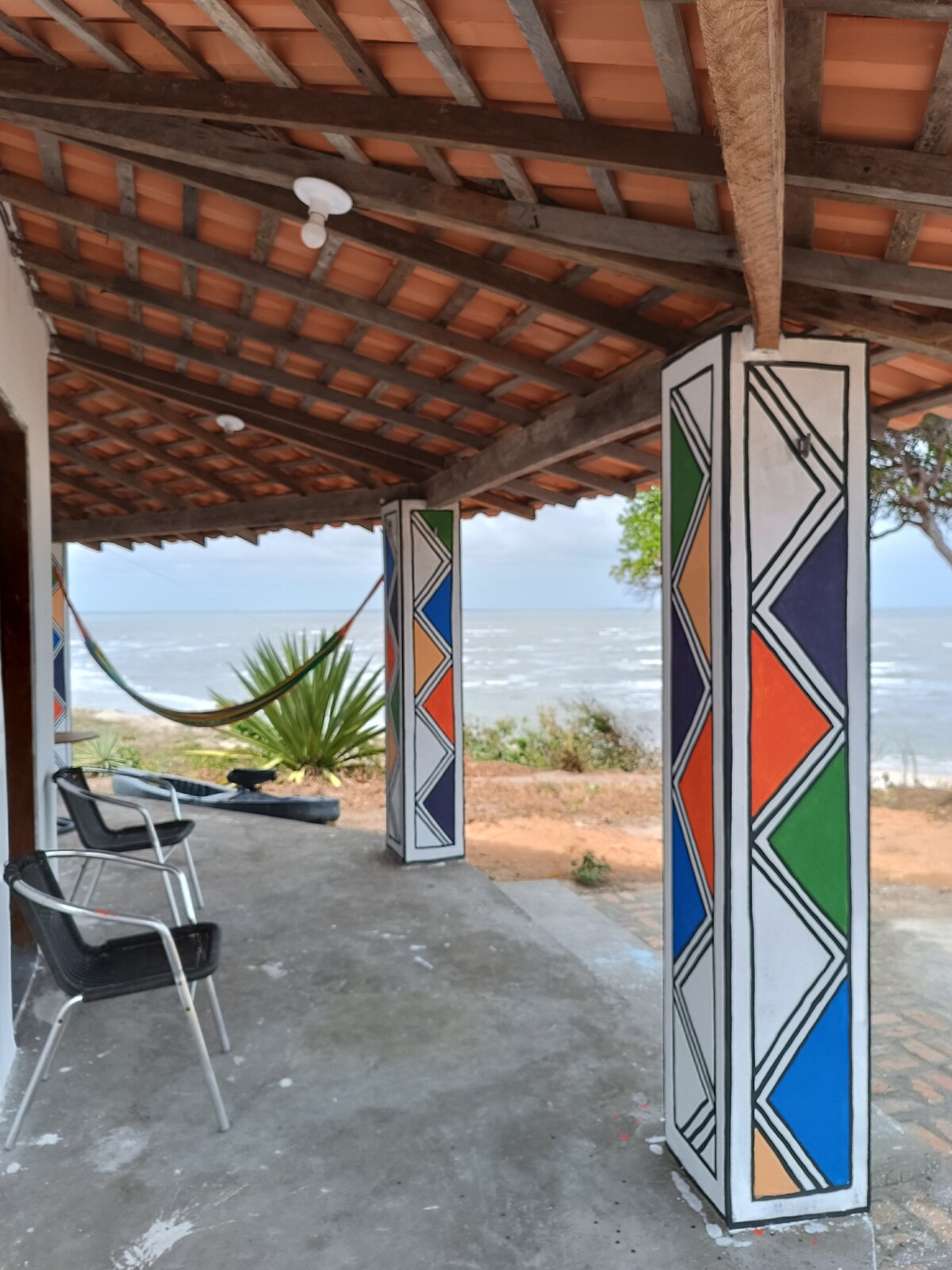A covered porch area features colorful, geometric columns adorned with vibrant patterns. Two black chairs are positioned on the concrete floor, while a hammock is visible in the background, adding to the relaxed atmosphere. The sea can be seen in the distance, enhancing the scenic view.