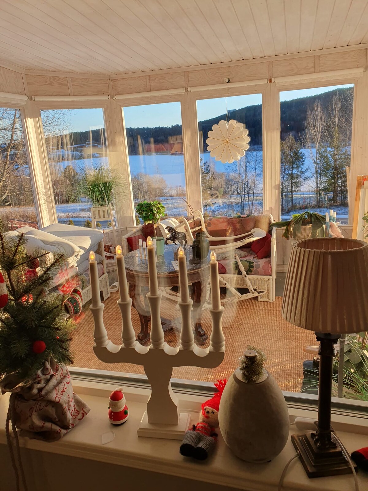 A sunlit sunroom features large glass windows offering a view of a winter landscape. Indoor plants are positioned near the windows, and decorative elements like candles and figurines add a cozy touch. A warm atmosphere is created by natural light reflecting off the scenery.