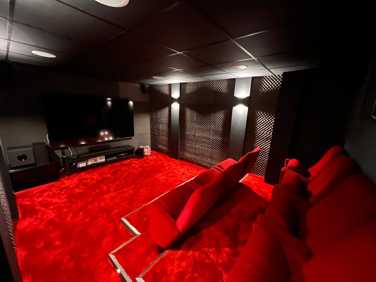 A private cinema room features plush red seating arranged in a semi-circle, facing a large flat-screen television. The room's walls are accented with textured panels, and the deep red carpet adds a cozy touch to the dark setting, enhancing the viewing experience.