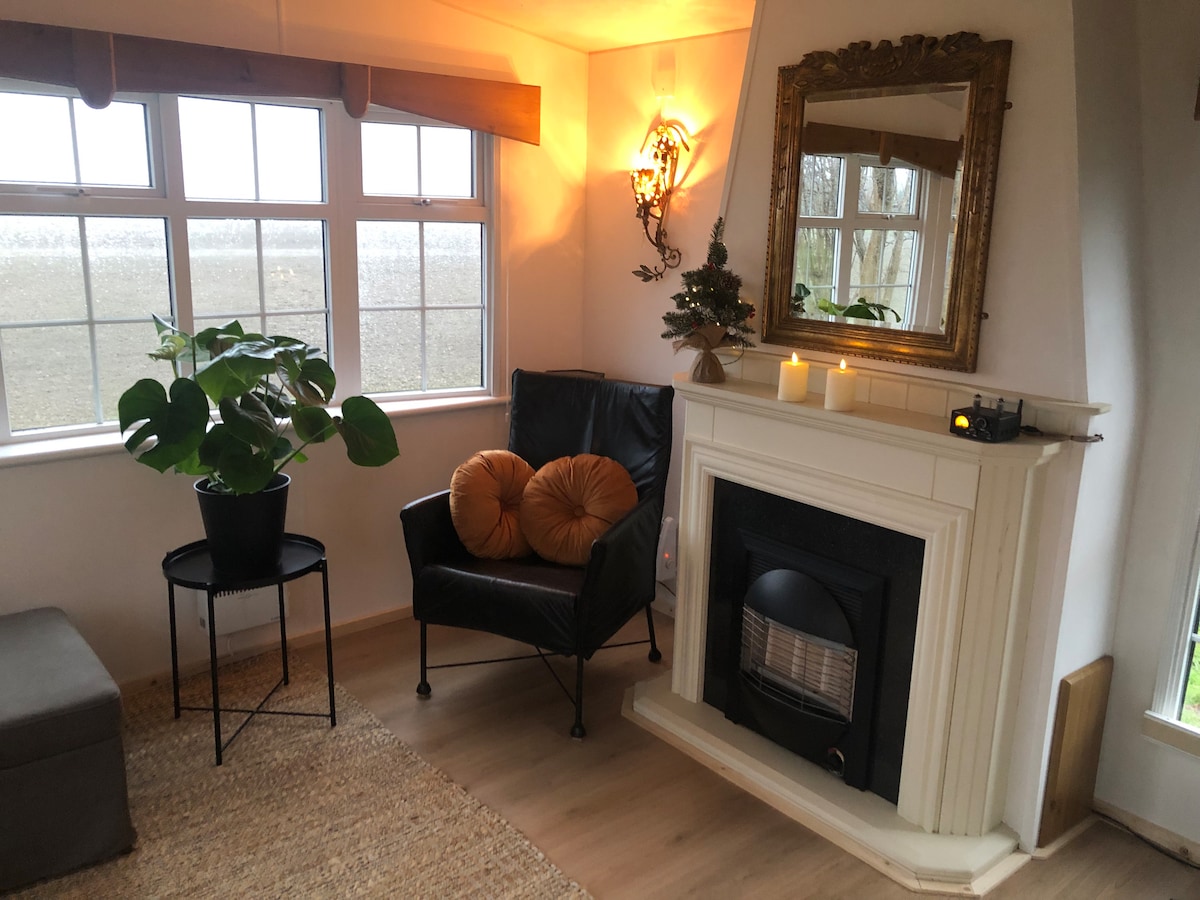 A corner of the living area features a black armchair with two orange cushions, positioned next to a small metal table. A decorative mirror hangs above a white fireplace, where candles are displayed, providing a soft glow. Large windows offer views of the surrounding landscape.