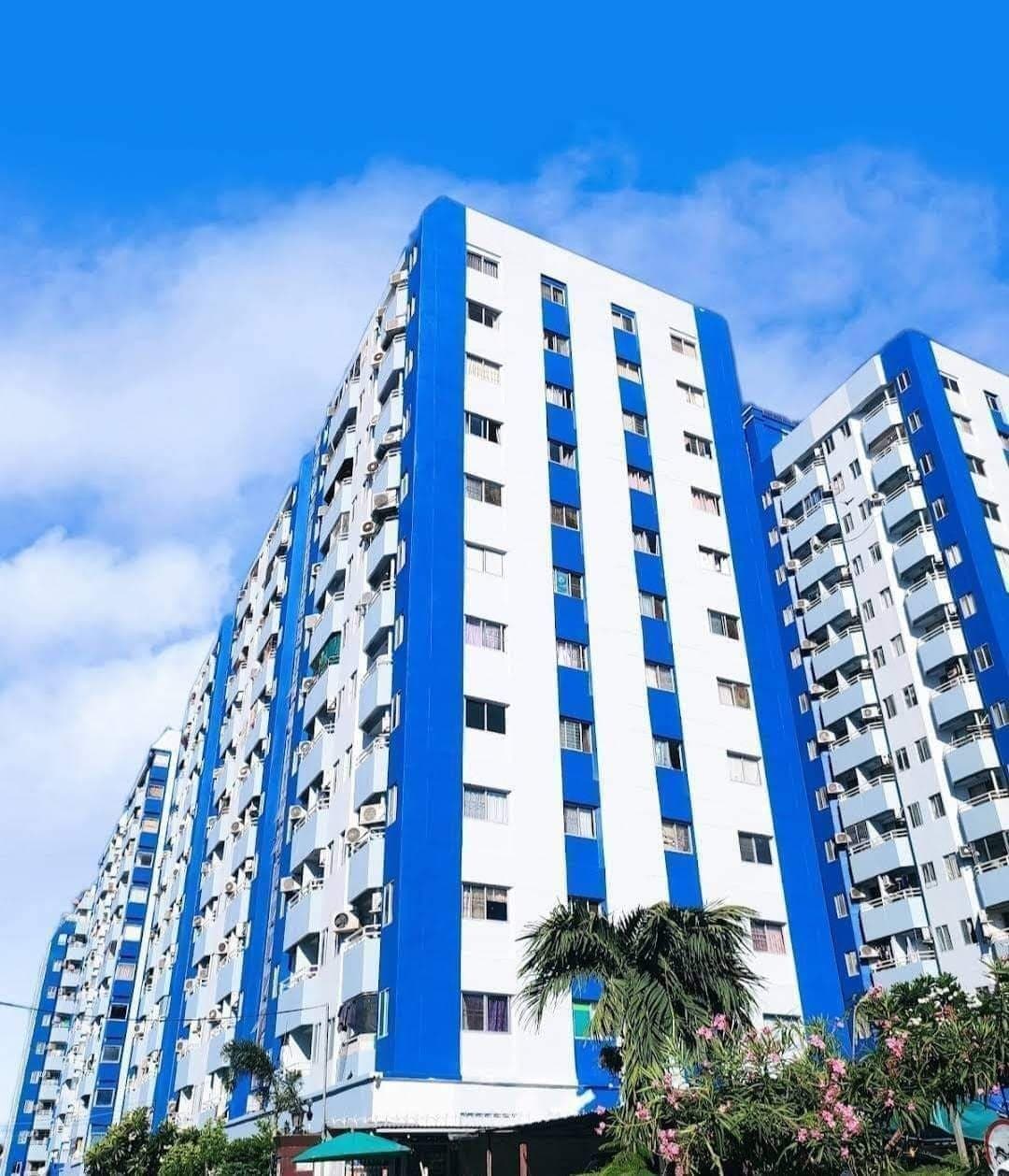 A modern multi-story building is displayed, featuring a combination of white and blue exterior walls. Numerous windows are visible, providing natural light to the units. Lush greenery surrounds the base of the structure, contributing to the overall inviting appearance.