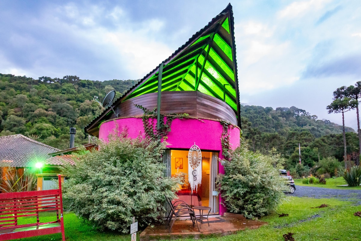A unique, multi-colored structure is framed by lush green vegetation. The architecture features a striking triangular roof with vibrant green highlights, and large windows offer glimpses of the interior. The pathway is lined with natural stone, leading to a welcoming entrance.