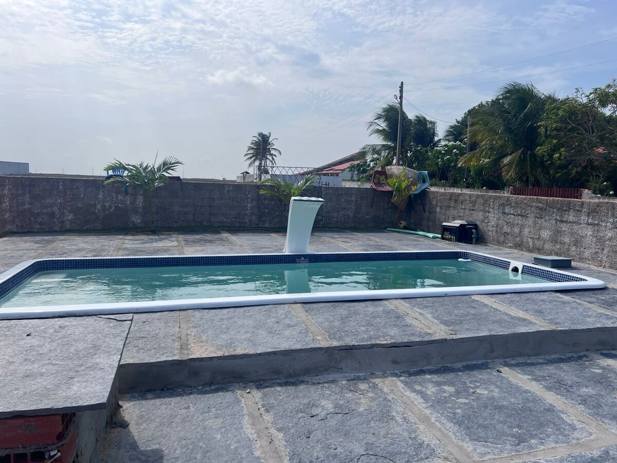 A serene outdoor area features a rectangular swimming pool surrounded by a concrete deck. Lush greenery is present along the edges, contributing to a relaxed atmosphere. Soft clouds are visible in the sky, hinting at a pleasant day.