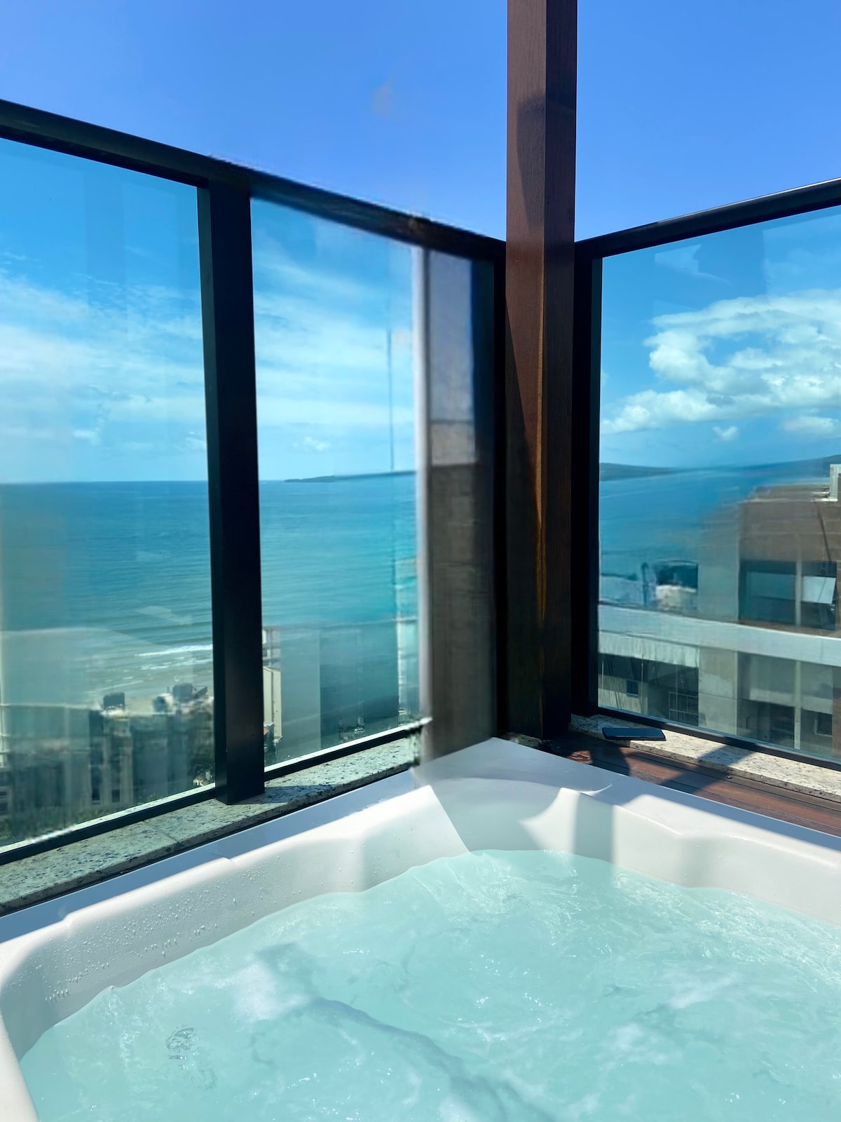A large window offers a panoramic view of the ocean and sky, with soft clouds visible. The jacuzzi, positioned beneath the window, is filled with water, inviting relaxation while enjoying the scenic coastal landscape.