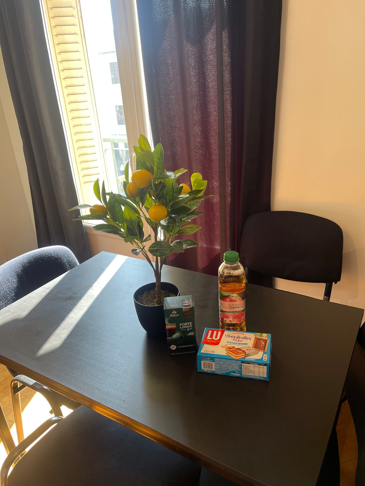 A small dining table is set with a potted lemon tree, a box of biscuits, and a bottle of beverage. Two chairs are positioned alongside the table. Natural light enters through a window adorned with dark curtains.