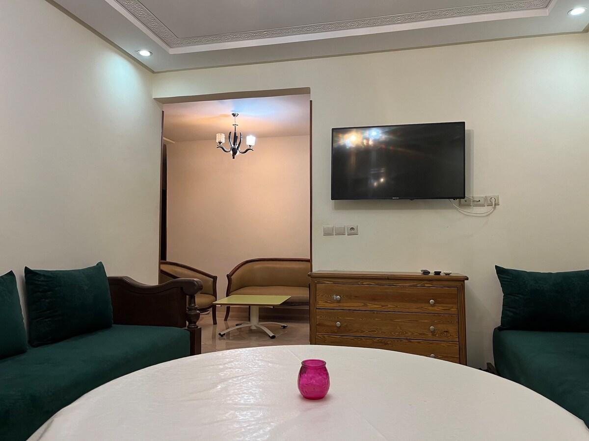 A living area features a round table at its center, complemented by green upholstered seating. A wooden dresser is positioned against the wall, while a flat-screen television is mounted above it. Soft lighting creates a cozy ambiance throughout the space.
