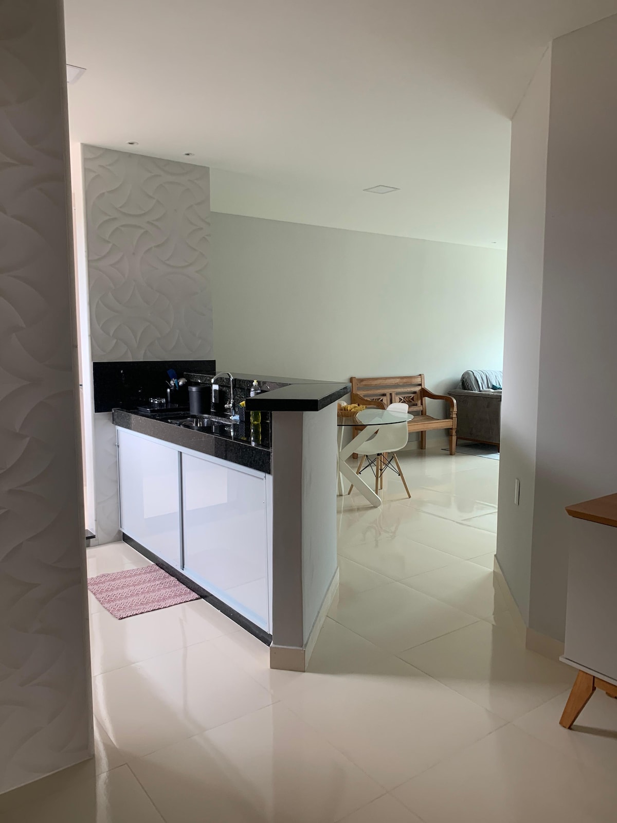 A modern kitchen area is visible, featuring sleek black countertops and white cabinetry. A pink mat lies on the floor in front of the countertop. In the background, a cozy living area is indicated, showcasing a brown sofa and a wooden chair.