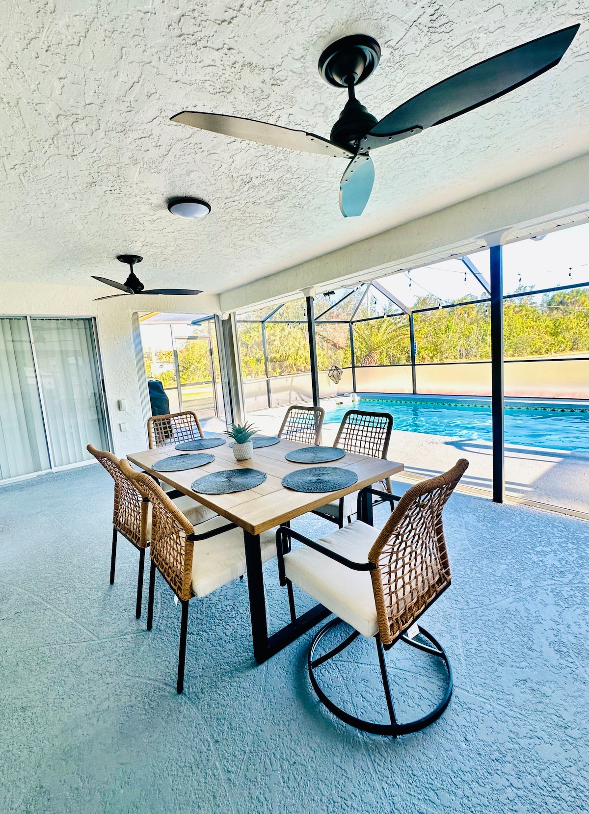A covered outdoor dining area is highlighted by a wooden table surrounded by six woven chairs. Two ceiling fans provide airflow. Large screen panels offer unobstructed views of a private pool and landscaped surroundings, ensuring a bright and inviting atmosphere.
