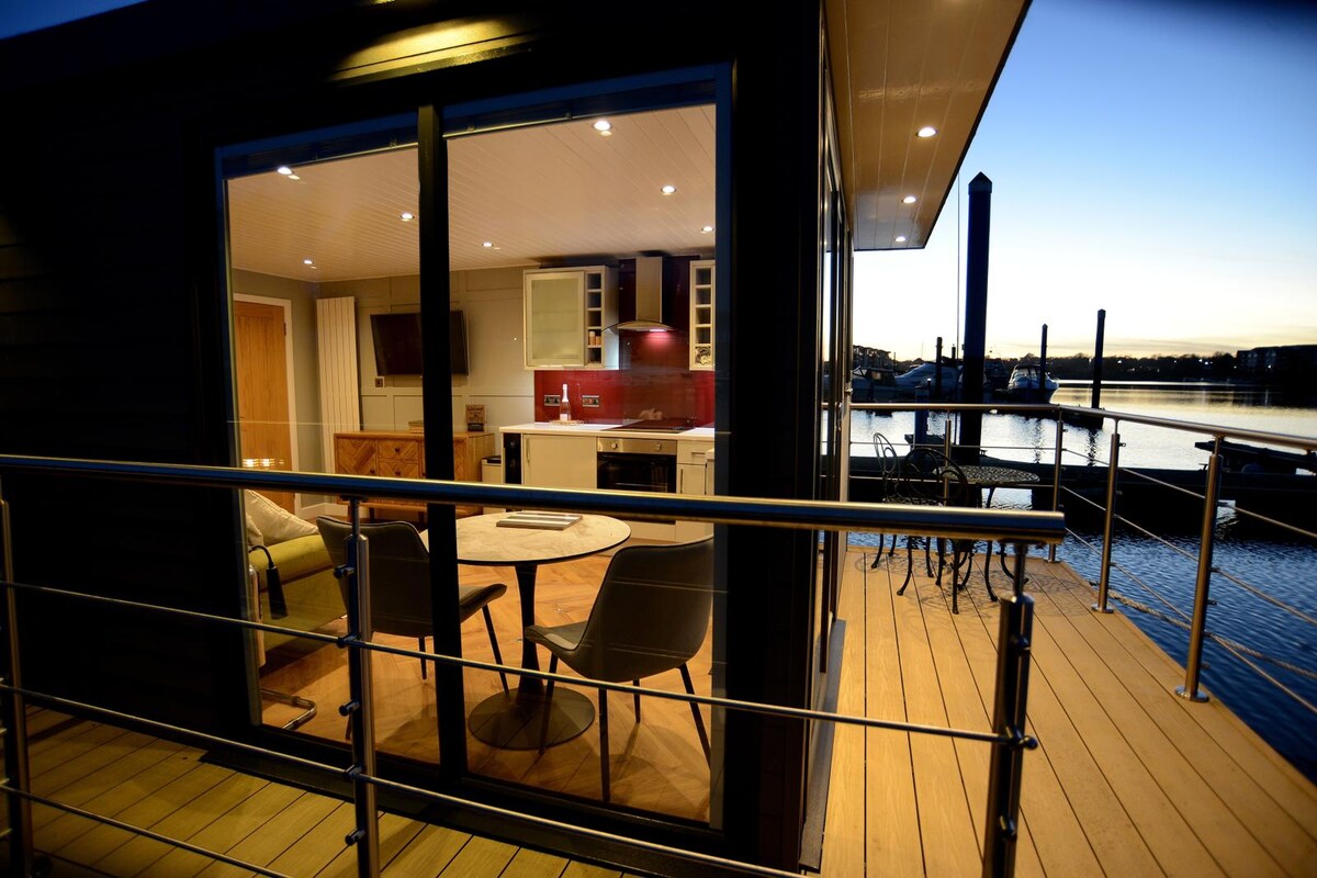 A spacious deck area is visible, leading to large glass doors that showcase the modern kitchen and seating area inside. The warm interior lighting highlights the contemporary design as it reflects against the water, creating an inviting ambiance for relaxing moments.