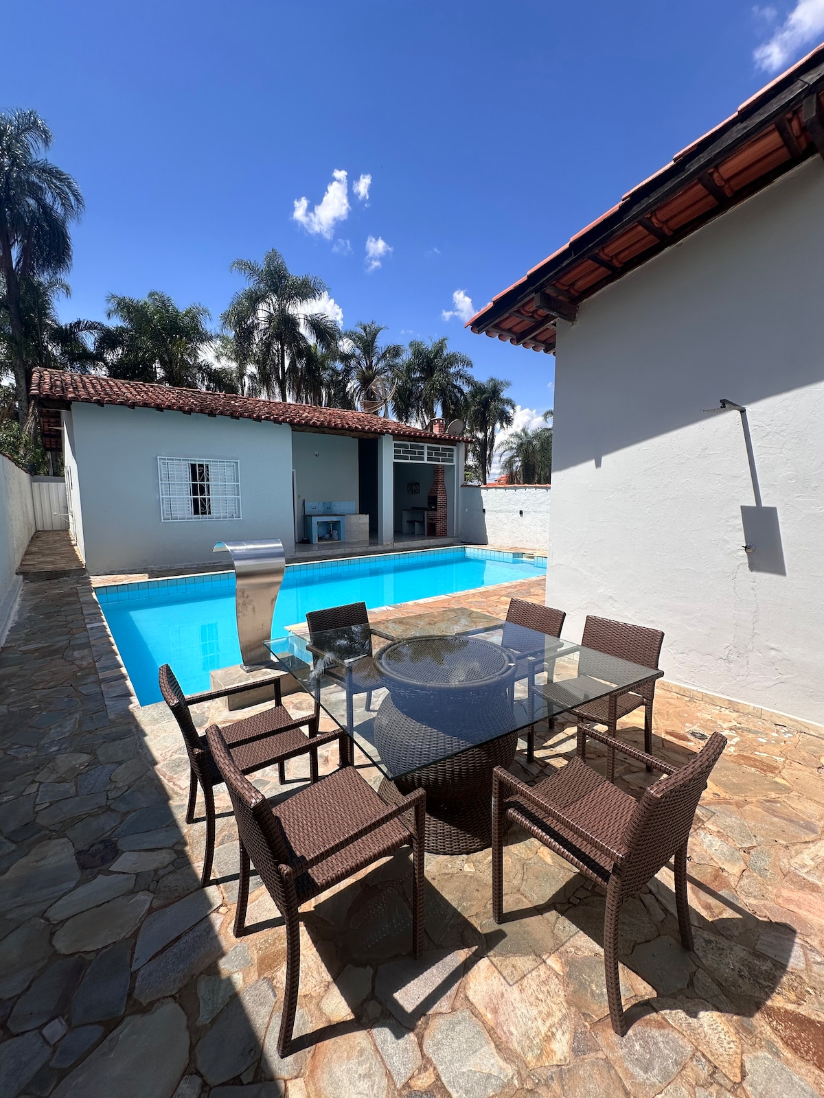 An outdoor dining area features a round glass-top table surrounded by six wicker chairs. A private pool, with crystal-clear water, reflects the surrounding palm trees and blue sky. The building in the background showcases light-colored walls and a red-tiled roof.