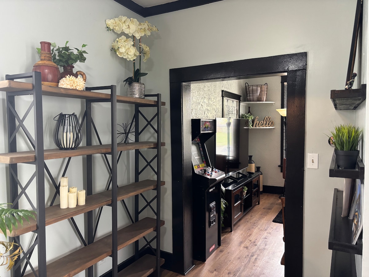 A cozy entryway is framed by dark wooden accents, featuring a stylish shelving unit adorned with decorative plants and candles. An arcade game and entertainment unit are visible through the doorway, enhancing the welcoming atmosphere of the living space.