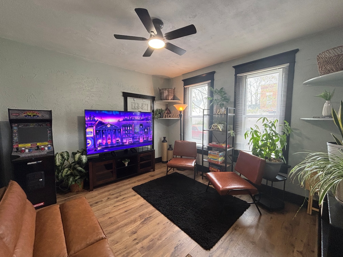 The living room features a large television positioned on a dark wood entertainment unit. Two brown chairs and a brown sofa create a cozy seating area. Plants are displayed near the windows, which allow natural light to brighten the room. A decorative rug covers the hardwood floor.