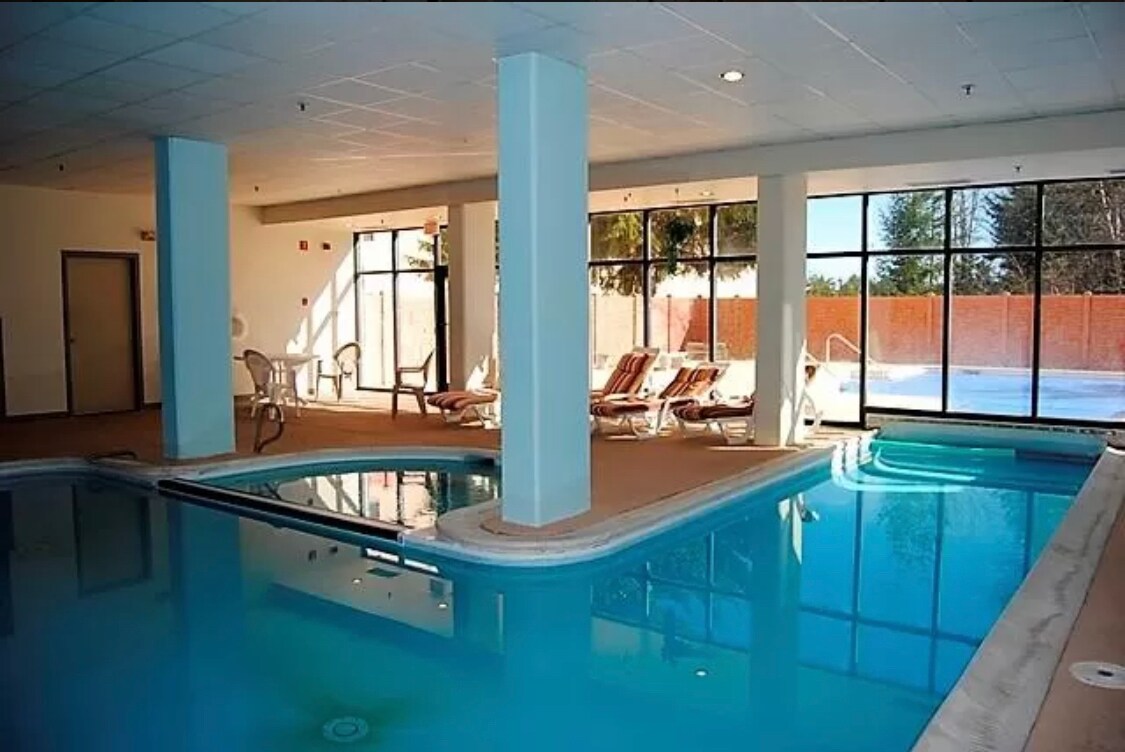 An indoor pool area features crystal-clear water with a hot tub situated nearby. Sunlight filters through large windows that overlook a patio space with lounge chairs. The ambiance is serene, inviting relaxation and enjoyment of the surrounding views.