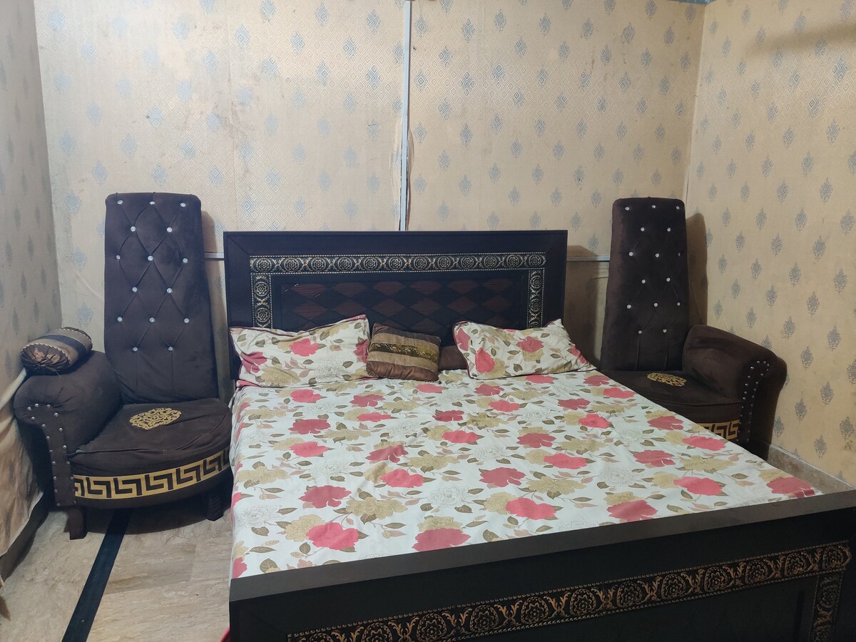 A sleeping area features a spacious bed adorned with floral bedding. Two upholstered chairs with decorative accents are positioned beside the bed, against a patterned wall. Natural lighting fills the room, enhancing the comfort of the space.