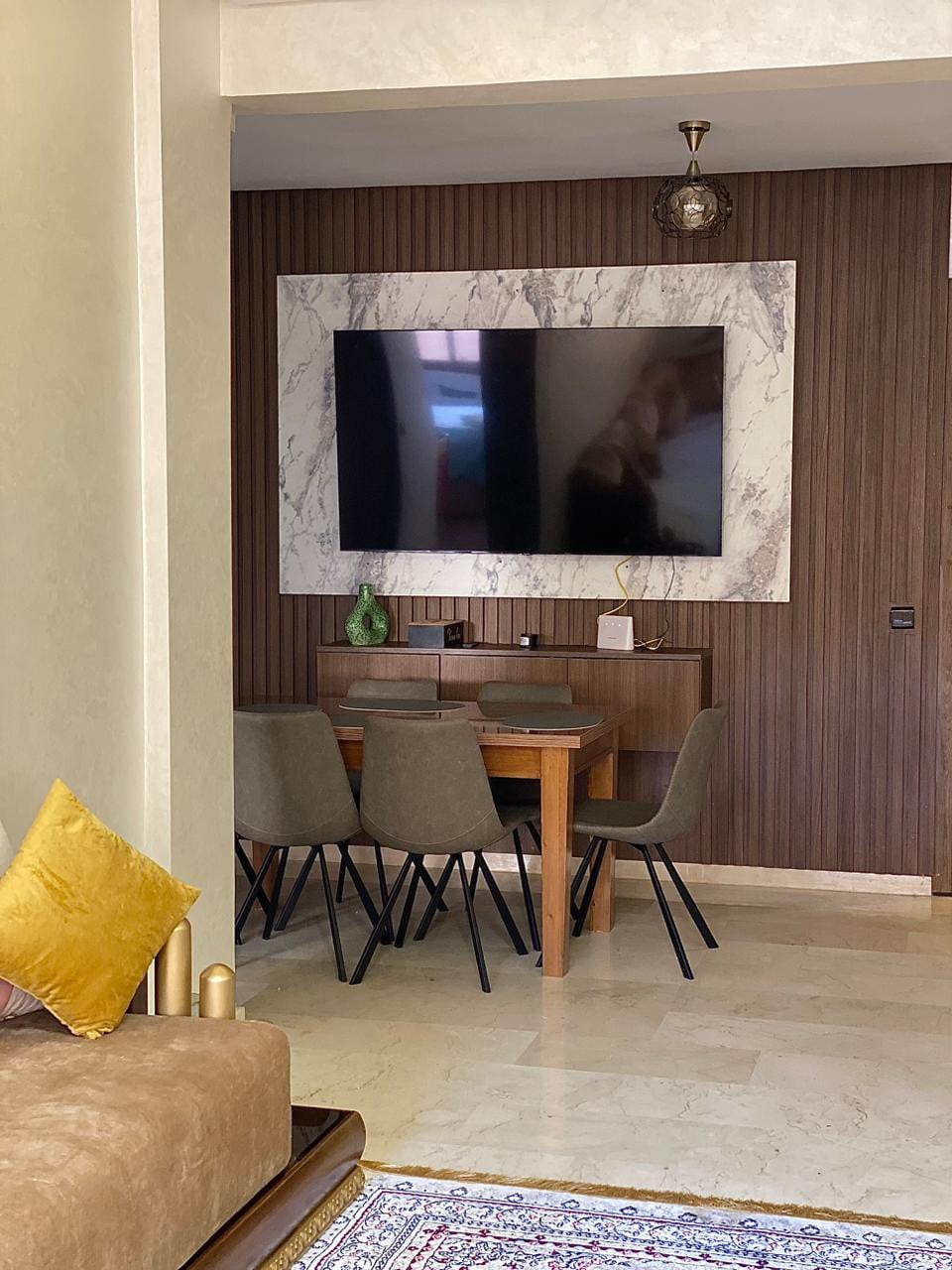 A dining area is visible, featuring a wooden table surrounded by four upholstered chairs. A large flat-screen TV is mounted on a textured wall with marble accents, and a green decorative item is displayed on the table. Soft lighting adds to the ambiance.