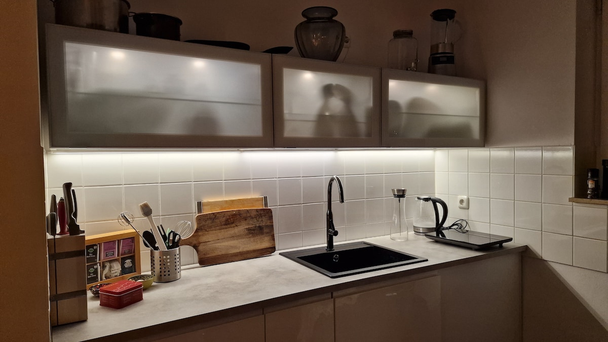The kitchen features modern cabinetry with frosted glass doors, reflecting soft lighting beneath the upper shelves. A sleek black sink is positioned next to wooden cutting boards, a coffee maker, and other cooking essentials on the countertop.