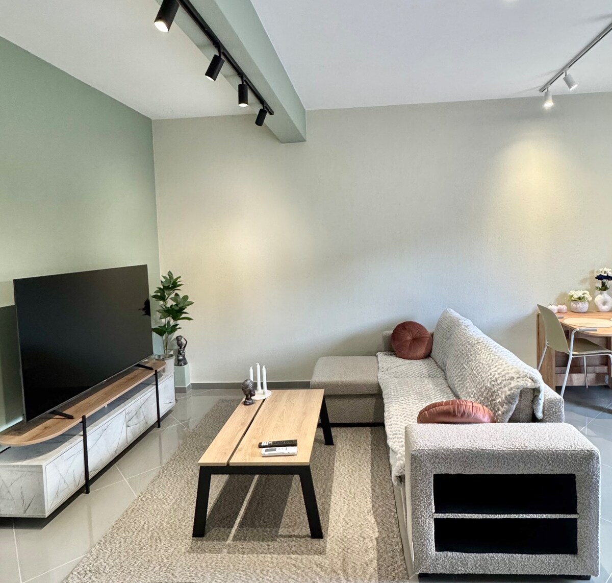 A spacious living area features a comfortable sofa against a light green wall. A modern television is positioned on a stylish stand, complemented by a decorative plant nearby. A wooden coffee table sits centrally on a soft area rug, and dining space is visible in the background.