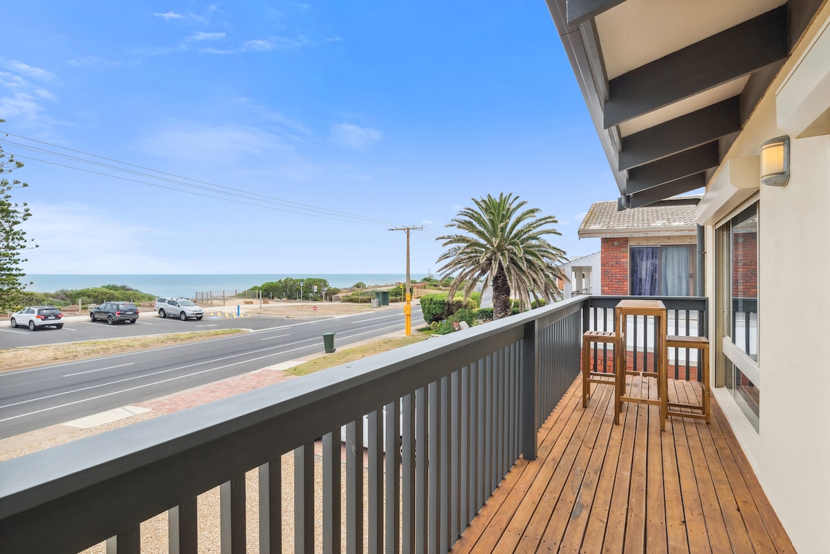 A spacious balcony is presented, featuring a wooden deck and two high-top chairs overlooking the beach. A clear view of the ocean and palm trees is visible, with the road lined by vehicles in the foreground, creating a serene outdoor setting.