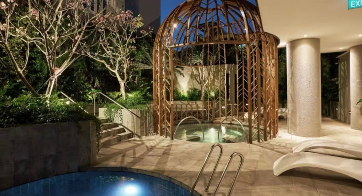 An outdoor area features a wooden structure surrounded by greenery and decorative trees. A small circular pool is visible alongside pathways, with lounge chairs positioned nearby. Soft lighting highlights the natural ambiance, creating a serene retreat.